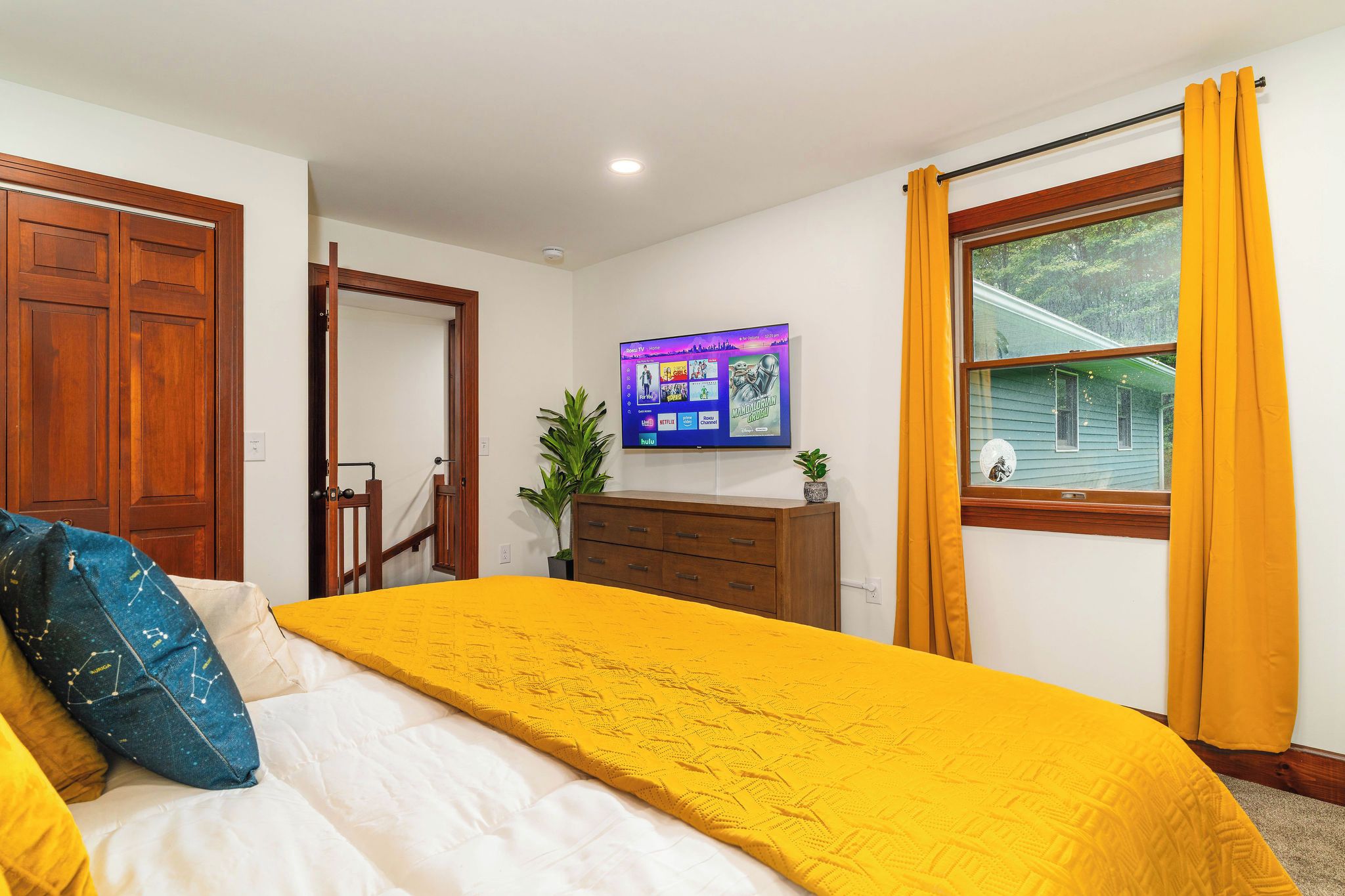 A bedroom with a bed, a dresser, a TV on the wall, and a window with curtains.