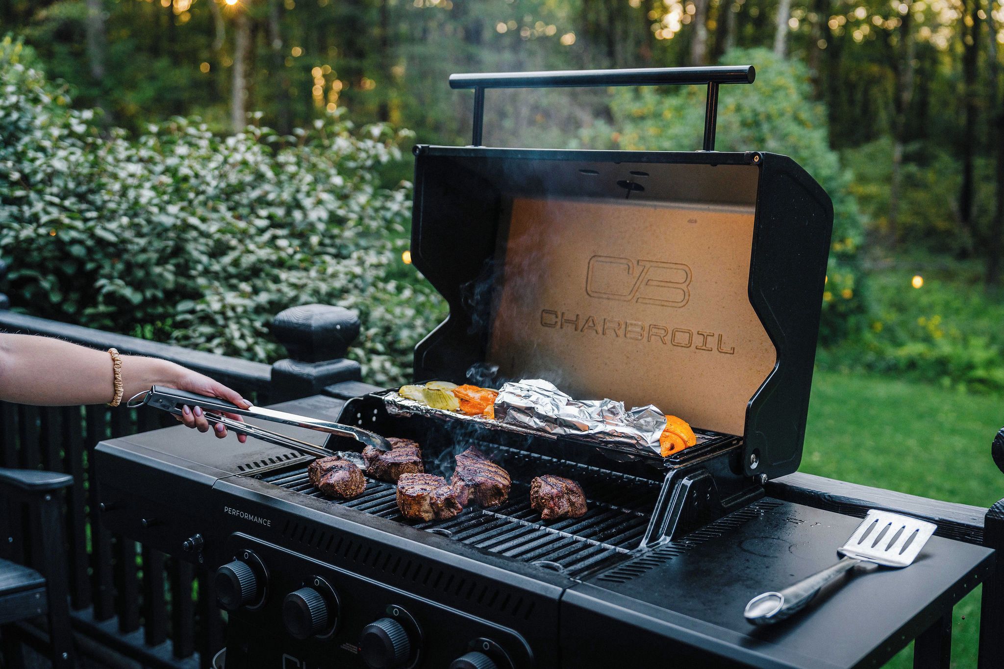 Un barbecue au charbon de bois avec plusieurs steaks en train de cuire, une spatule et des aliments enveloppés dans du papier d’aluminium sur le gril.