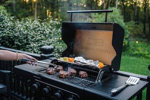 Una parrilla de carbón con varios filetes asándose, una espátula y alimentos envueltos en papel de aluminio sobre la parrilla.