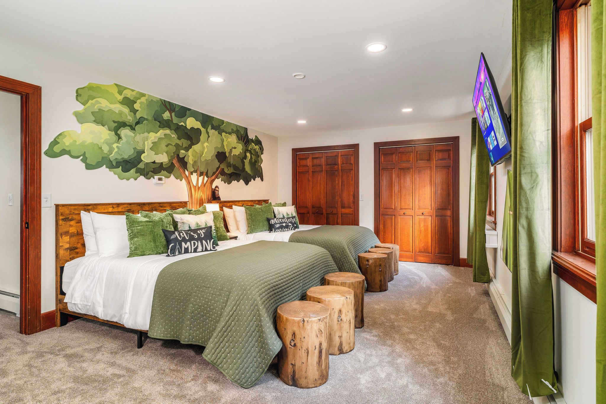 A bedroom with a large bed, two bedside tables, a wall-mounted TV, and a large tree mural.