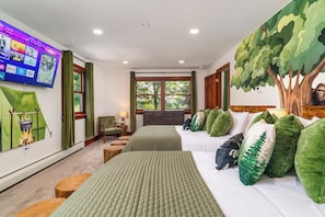 Un dormitorio con un televisor montado en la pared mostrando varios canales, una cama con ropa de cama verde y blanca, una silla y una ventana con vistas a árboles.