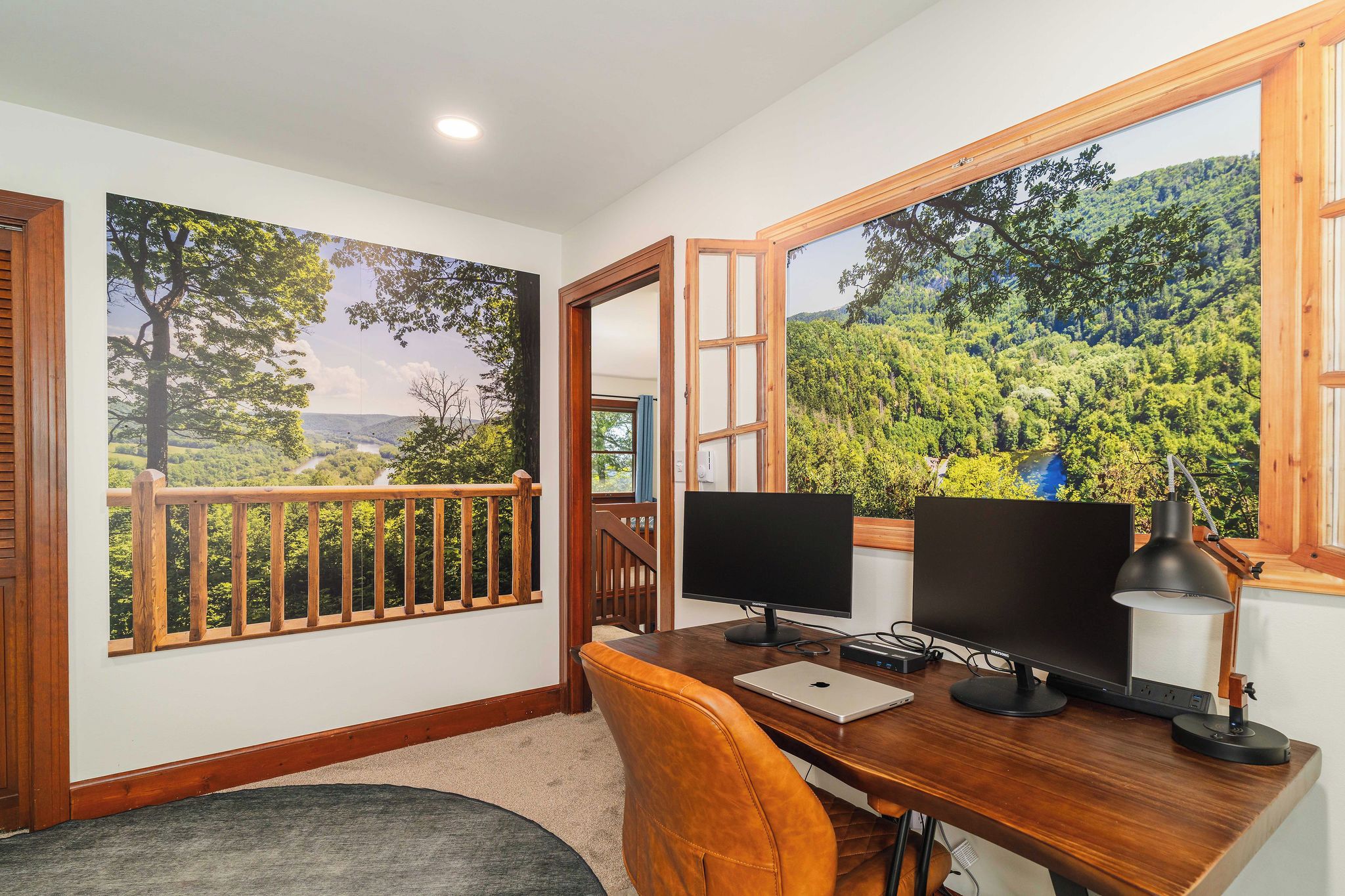 Une pièce comprenant un bureau, deux écrans d’ordinateur, un ordinateur portable et une lampe de bureau. Une fenêtre offre une vue sur une forêt et une rivière.
