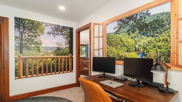 A room with a desk, two monitors, a laptop, and a desk lamp. There is a window with a view of a forest and a river.