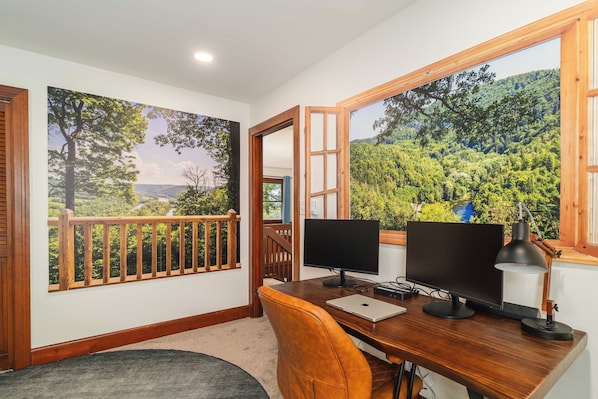 Una habitación con un escritorio, dos monitores, una computadora portátil y una lámpara de escritorio. Hay una ventana que ofrece vistas a un bosque y un río.