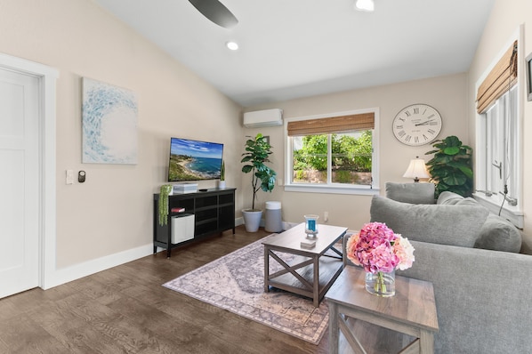 A living room with a sofa, a coffee table with a vase of flowers, a television, a clock, and a window with a view of greenery.