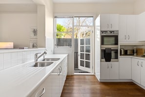 A modern kitchen with white cabinetry, stainless steel appliances, and a view through an open door to the outdoors. - Waterfall Garden 3-Bed Home McMahons Point (McMahons Point)