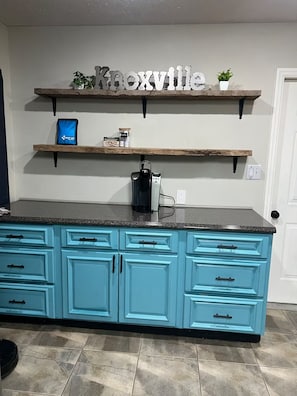 A kitchen with a teal cabinetry, a coffee maker on the counter, and a shelf with a sign above. - West Knox home w/Hot Tub and Fenced Backyard (Knoxville)