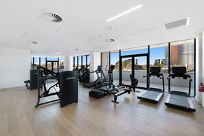 Academia moderna com diversos equipamentos de exercício, amplas janelas e vista para edifícios.