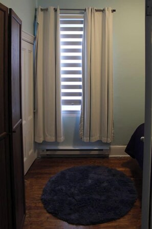 Una habitación con ventana, persianas horizontales, una alfombra azul oscuro en el suelo y una puerta a la izquierda.