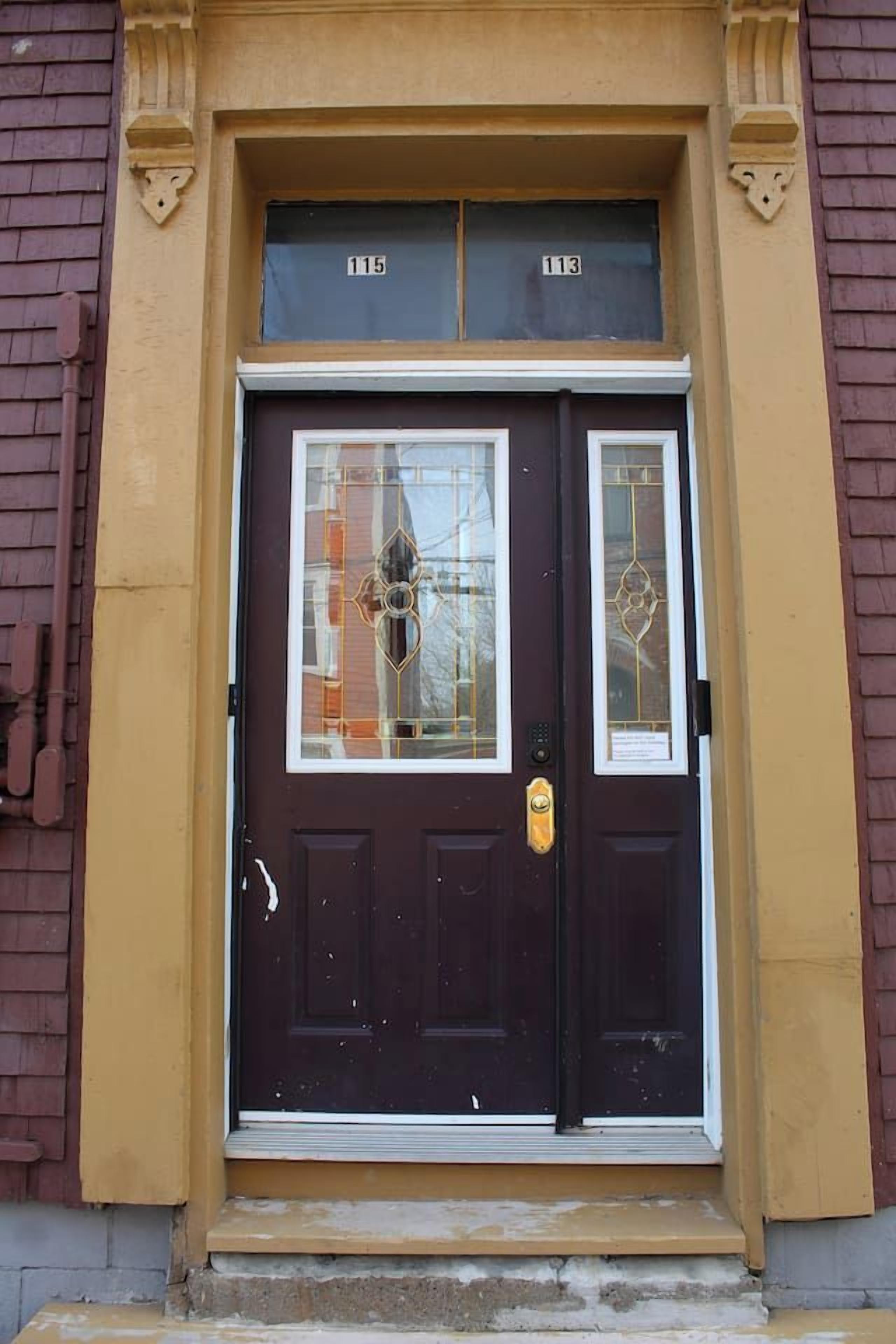 Une porte à deux battants de couleur brun foncé, avec un panneau de verre et une petite fenêtre au-dessus, intégrée à la façade jaune d’un immeuble.