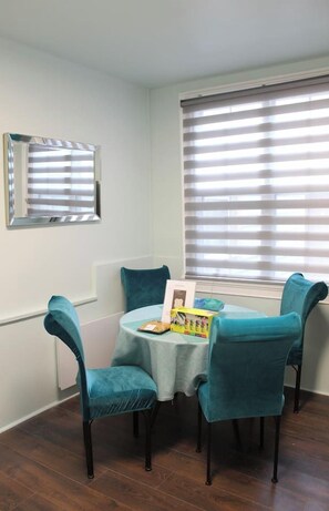 A dining area with a round table, four teal chairs, a mirror on the wall, and a window with blinds. - Uptown Saint John Retreat (Saint John)