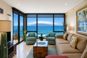 A spacious living room with a large window overlooking the ocean, a comfortable sofa, two armchairs, a coffee table, and a decorative lamp. - Oceanfront Mahana Ka'anapali | AC, Pool, Hot Tub, Pickleball, King Bed (Lahaina)