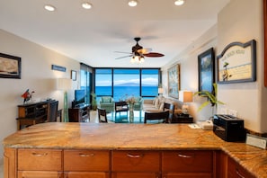 A spacious living area with a large window overlooking the ocean, a ceiling fan, and a kitchen area with a granite countertop. - Oceanfront Mahana Ka'anapali | AC, Pool, Hot Tub, Pickleball, King Bed (Lahaina)