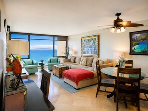 A spacious living area with a large window offering a view of the ocean, a ceiling fan, and a dining table with chairs. - Oceanfront Mahana Ka'anapali | AC, Pool, Hot Tub, Pickleball, King Bed (Lahaina)