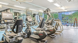 Ein Fitnessstudio mit verschiedenen Trainingsgeräten, einem großen Spiegel und Blick auf eine gebirgige Landschaft durch die Fenster.