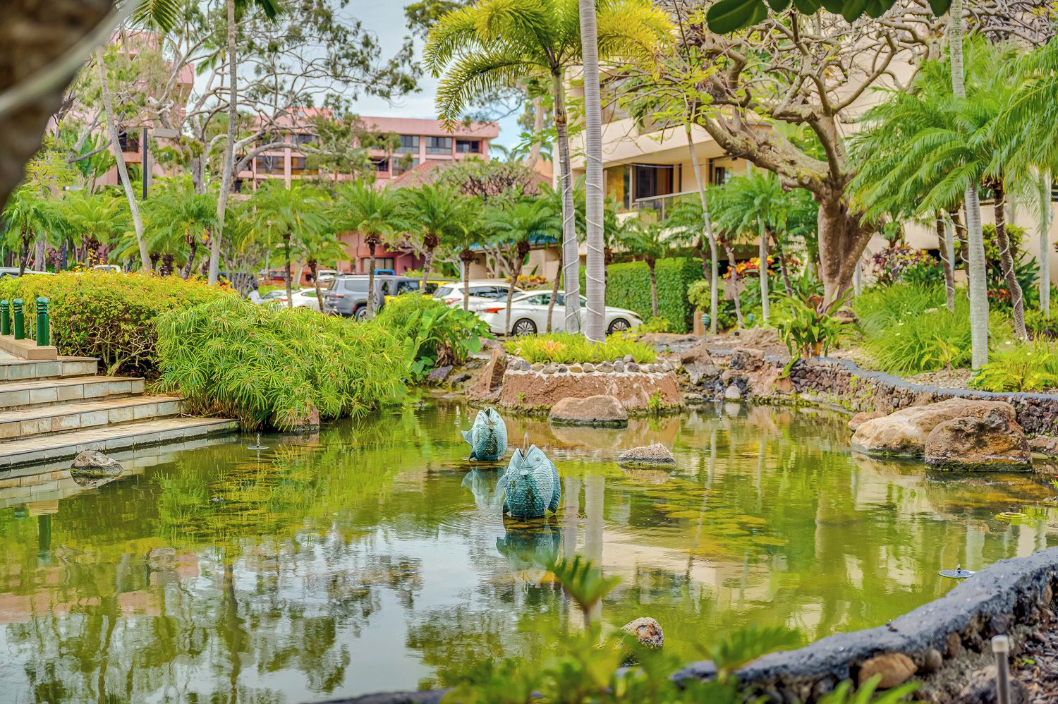 A serene pond with decorative fish sculptures, surrounded by lush greenery and a building in the background.