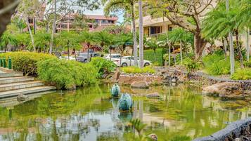 A serene pond with decorative fish sculptures, surrounded by lush greenery and a building in the background.