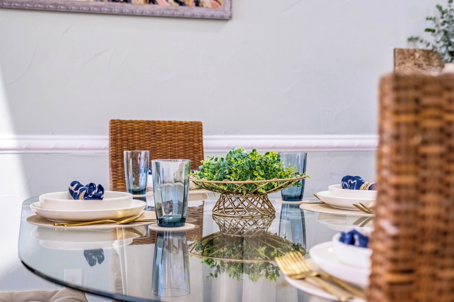 A dining table with clear glassware, white plates, and gold cutlery, set against a wicker chair and a wall-mounted picture frame.