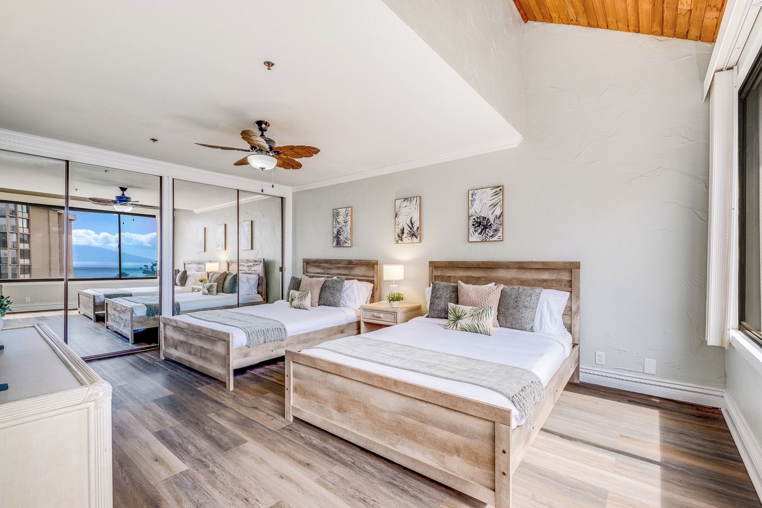 A modern bedroom with two beds, a ceiling fan, and a large window overlooking the ocean.