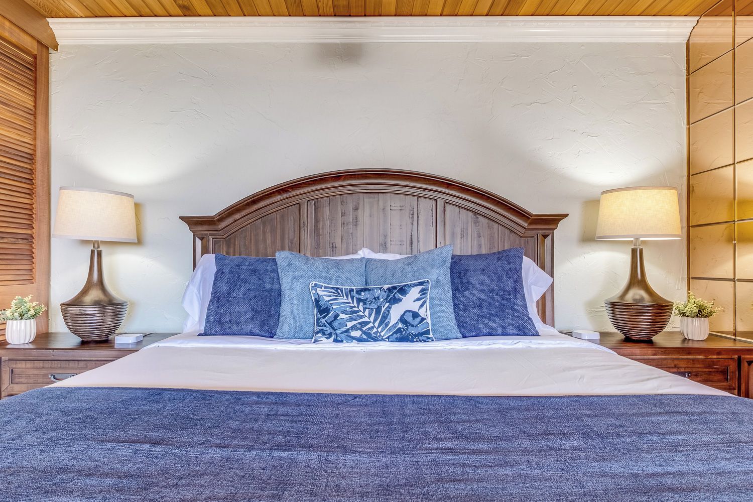 Een slaapkamer met een groot bed, een houten hoofdbord, twee nachtkastjes met lampen en een blauw geweven sprei.