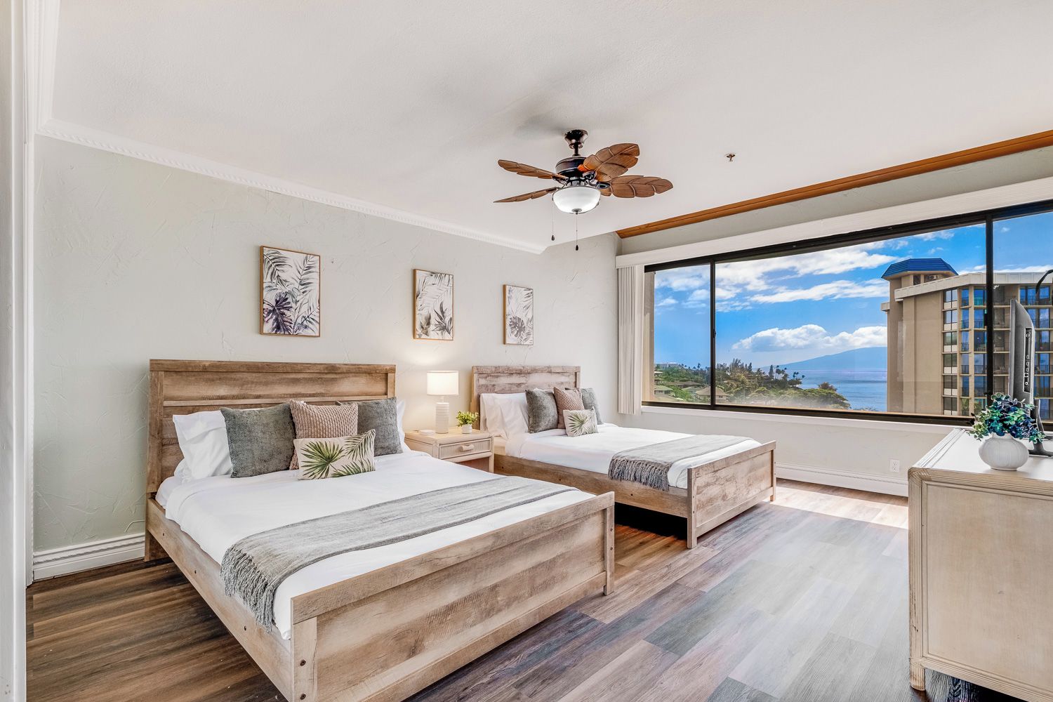 Une chambre à coucher avec deux lits en bois, un ventilateur de plafond et une grande fenêtre donnant sur un bâtiment et un paysage.