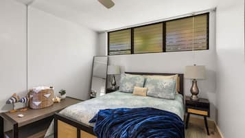 Ein Schlafzimmer mit einem Bett, zwei Nachttischen, einem kleinen Tisch, einem Deckenventilator und einem Fenster mit Jalousien.