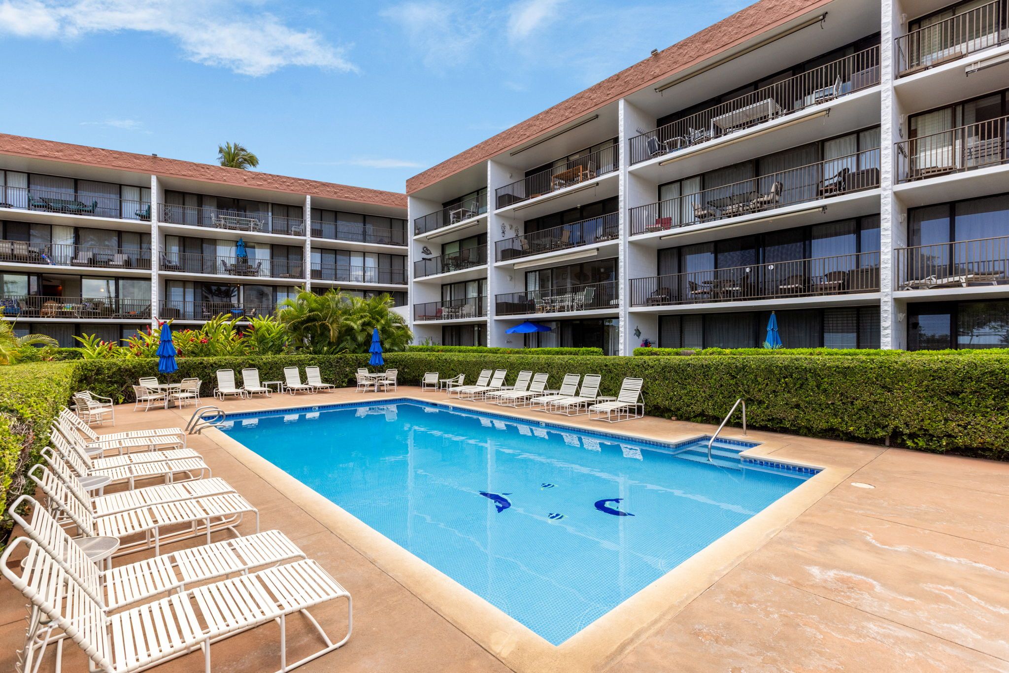 Ein Hotel mit Schwimmbad, umgeben von Liegestühlen und Sonnenschirmen, mehreren Balkonen und einem gepflegten Garten.