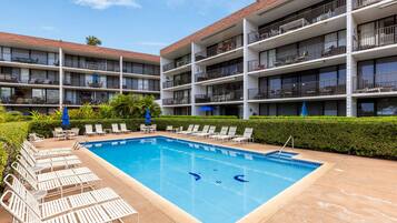 Ein Hotel mit Schwimmbad, umgeben von Liegestühlen und Sonnenschirmen, mehreren Balkonen und einem gepflegten Garten.