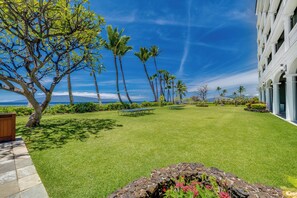 A well-maintained lawn with a stone wall and flower bed in the foreground, a building with a balcony on the right, and a clear view of the ocean and palm trees in the background.