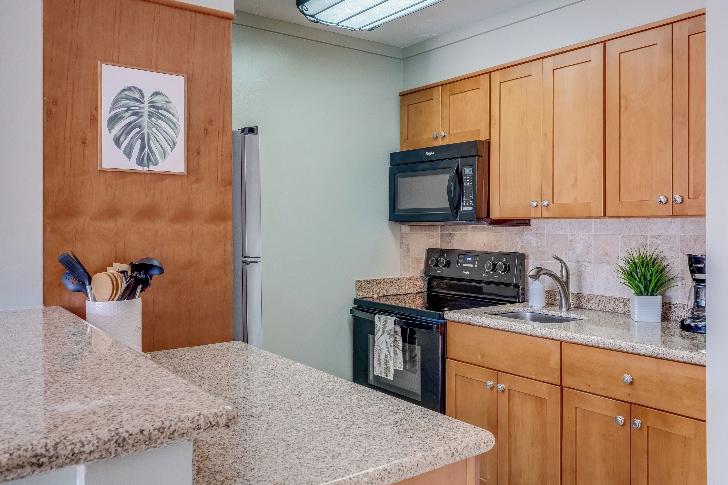 A kitchen with granite countertops, wooden cabinets, a stainless steel refrigerator, a black microwave, a stove, and a sink.