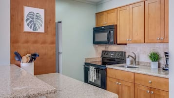A kitchen with granite countertops, wooden cabinets, a stainless steel refrigerator, a black microwave, a stove, and a sink.
