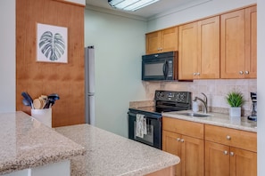 A kitchen with granite countertops, wooden cabinets, a stainless steel refrigerator, a black microwave, a stove, and a sink.