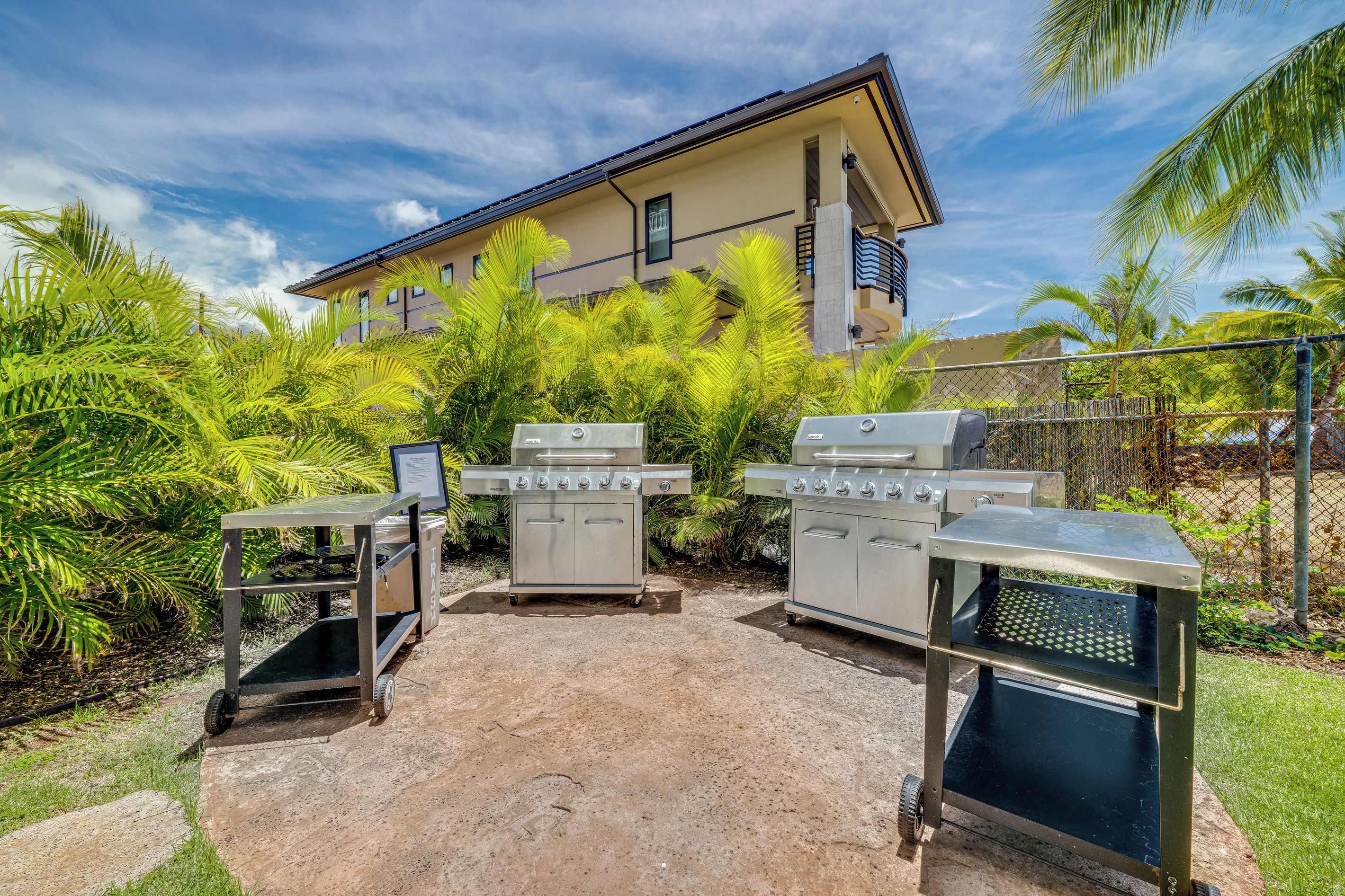 An outdoor barbecue area with a grill and a trash bin, surrounded by greenery and a building in the background.