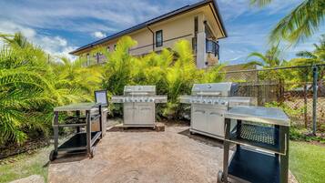 An outdoor barbecue area with a grill and a trash bin, surrounded by greenery and a building in the background.