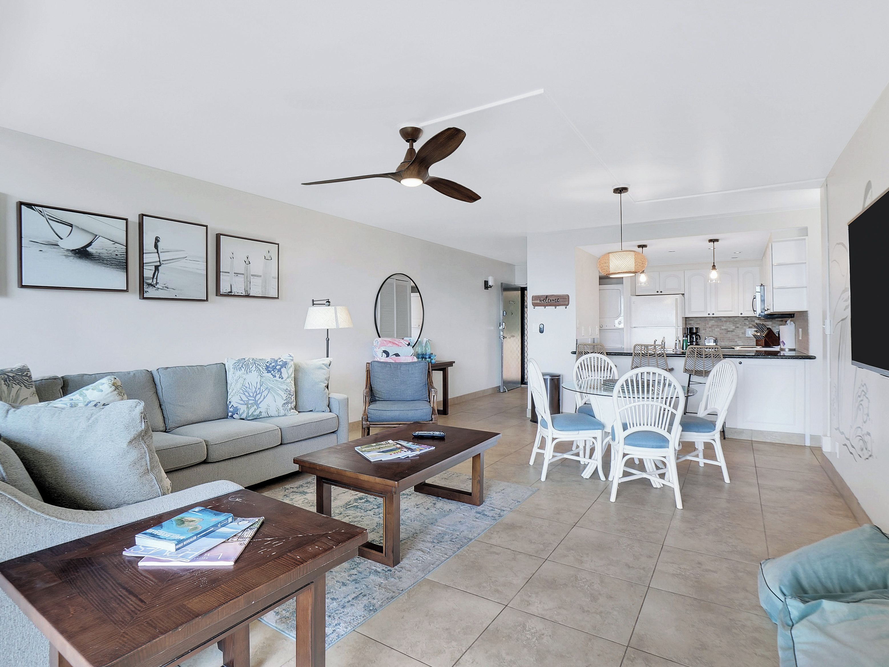 A modern living room with a sofa, coffee table, and dining area with chairs. There is a ceiling fan, a kitchen in the background, and artwork on the walls.