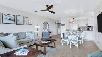 A modern living room with a sofa, coffee table, and dining area with chairs. There is a ceiling fan, a kitchen in the background, and artwork on the walls.