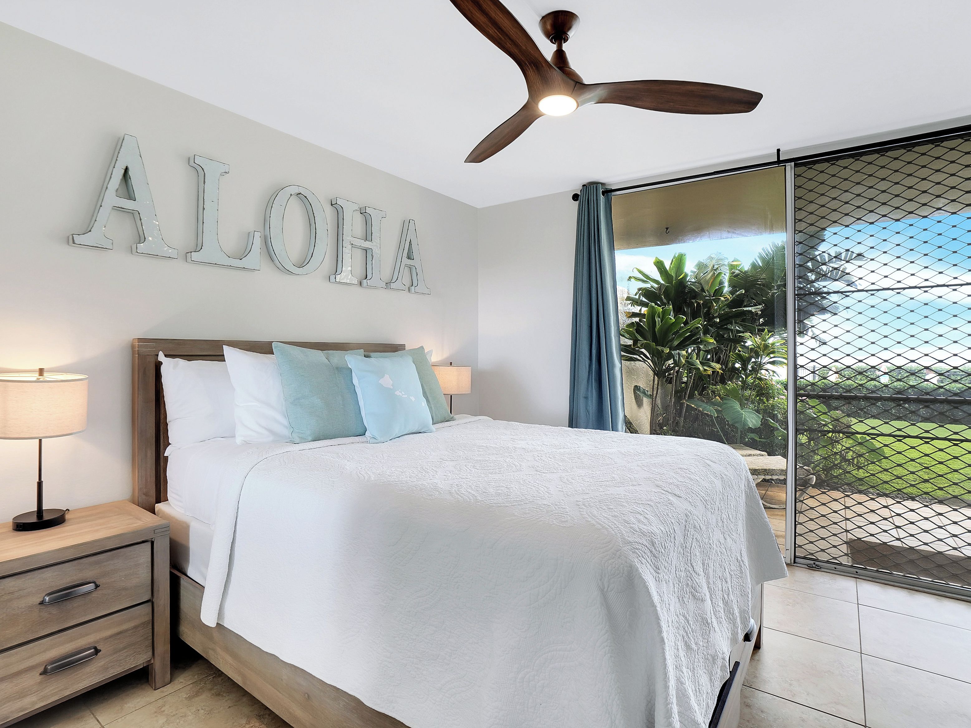 A bedroom with a bed, nightstands, a ceiling fan, and a view of greenery through a window.
