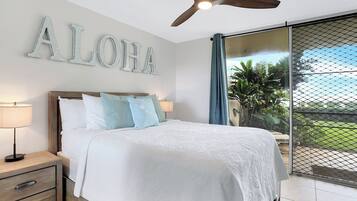 A bedroom with a bed, nightstands, a ceiling fan, and a view of greenery through a window.
