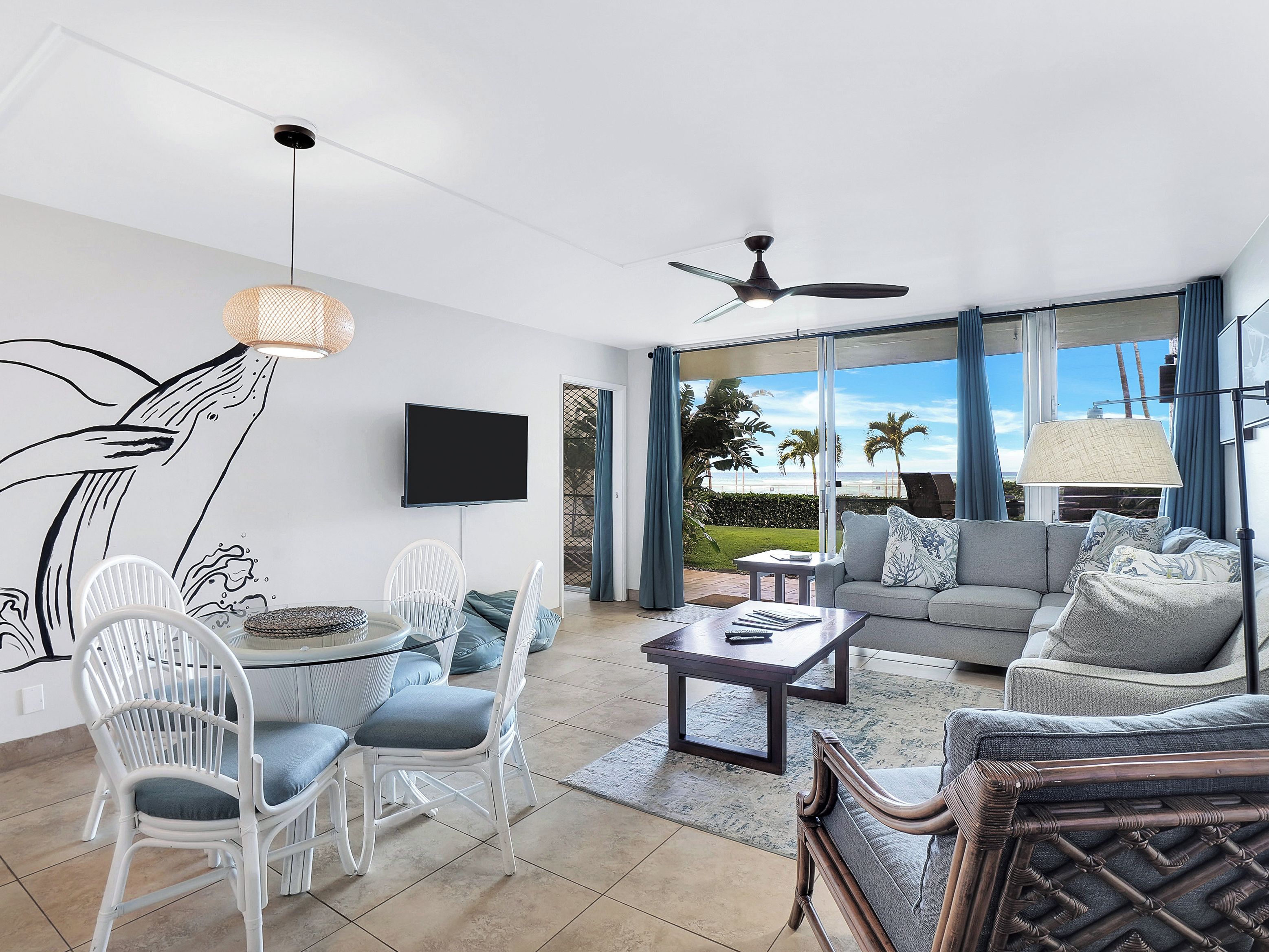 A modern living room with a flat-screen TV, white wicker chairs, a grey sofa, and a ceiling fan.