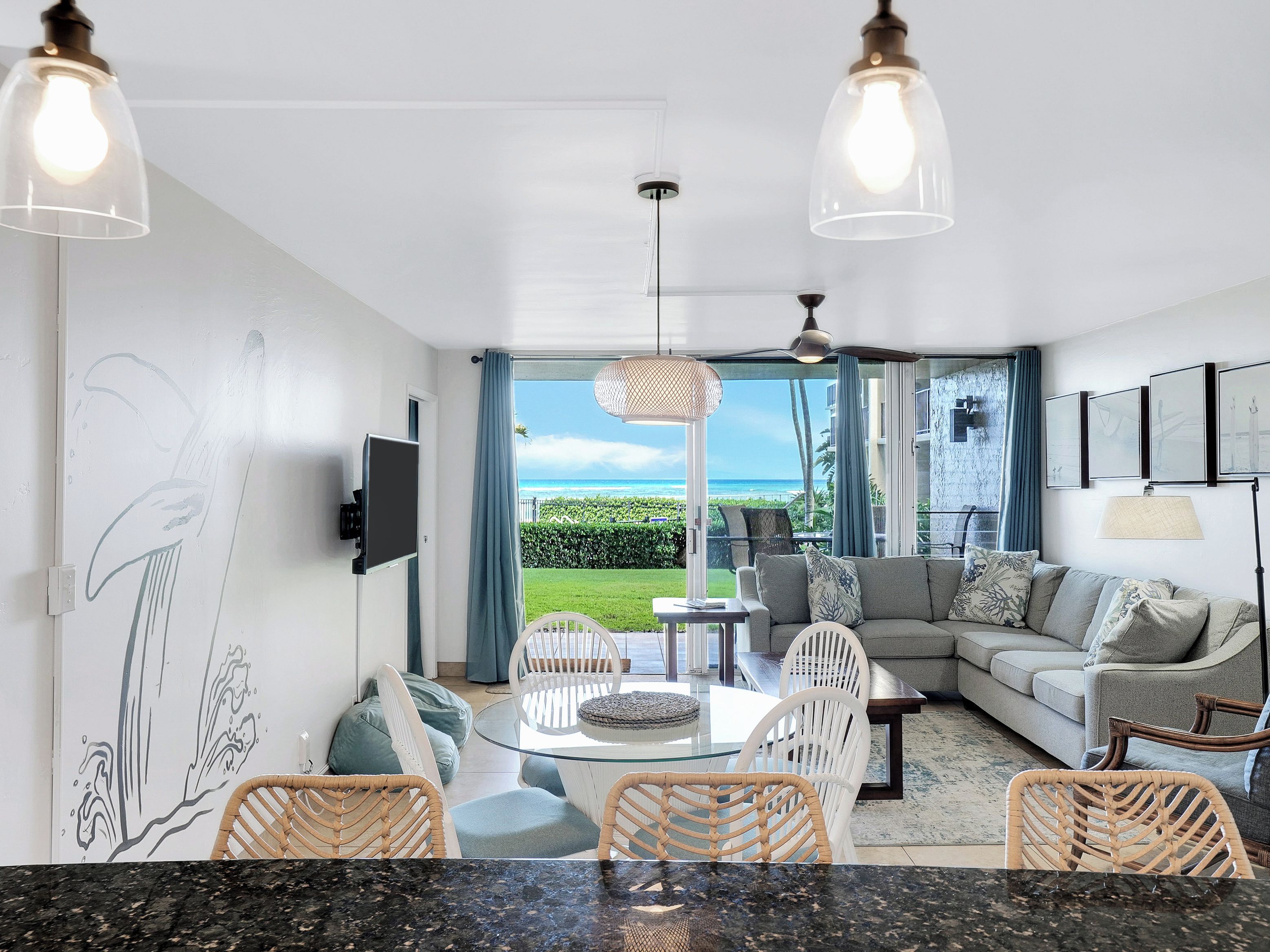 A modern living room with a glass dining table, wicker chairs, a grey sofa, and a view of the ocean.