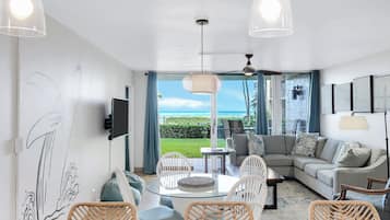 A modern living room with a glass dining table, wicker chairs, a grey sofa, and a view of the ocean.