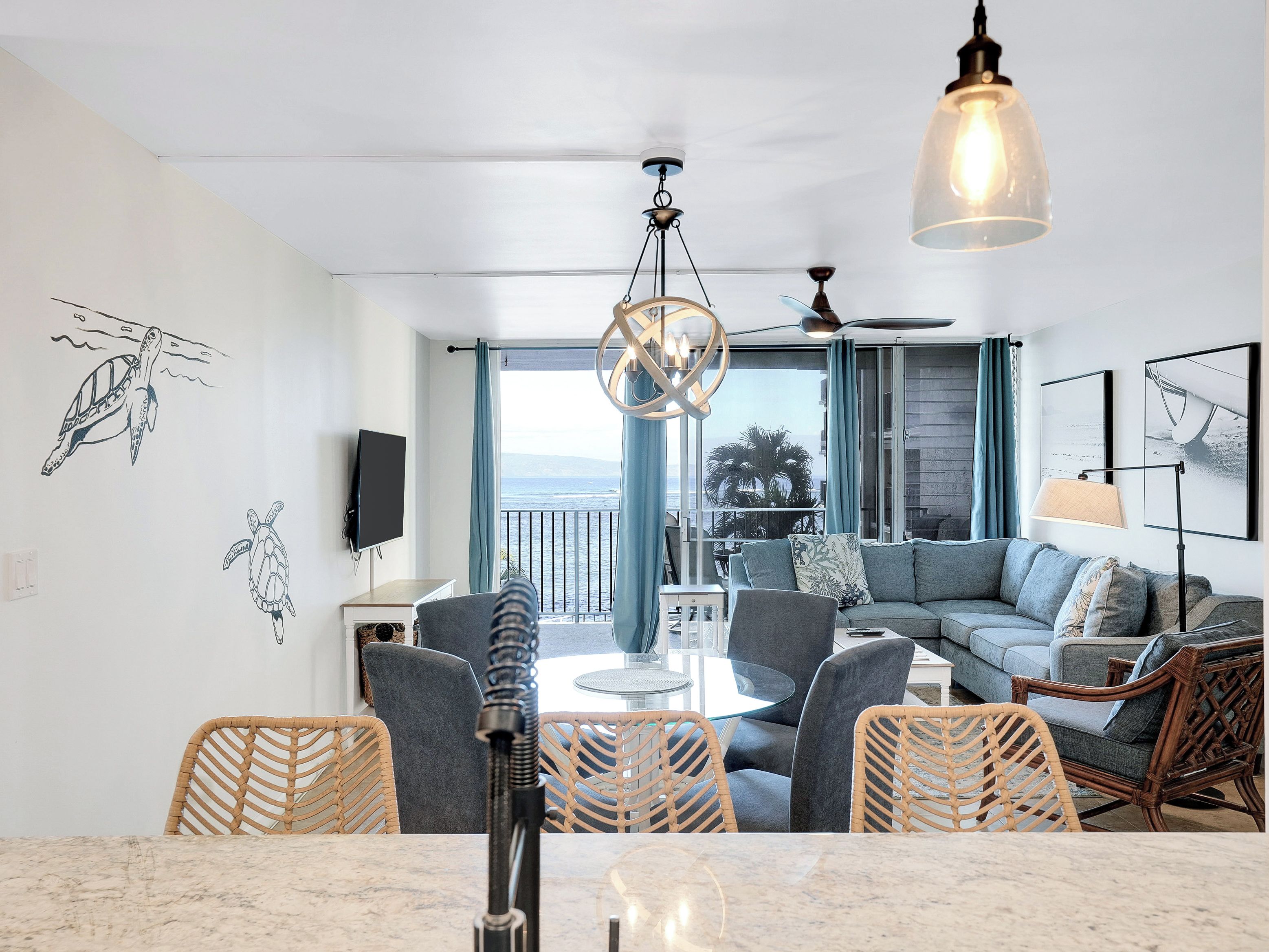 Una sala de estar moderna con un sofá grande, una mesa de comedor y sillas, un ventilador de techo y vista al océano a través de amplias ventanas.