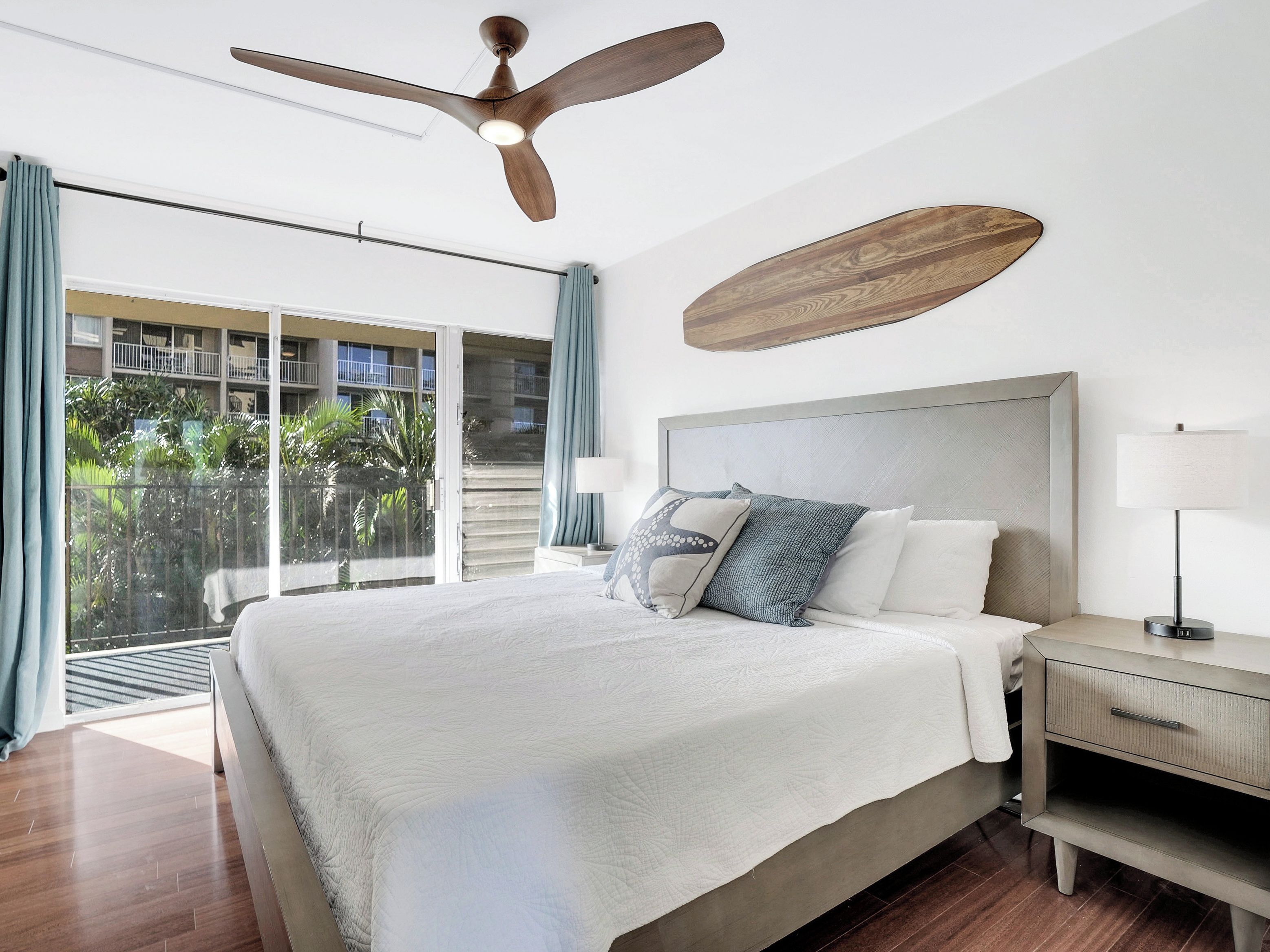 Une chambre à coucher comprenant un lit, une table de chevet, une lampe, un ventilateur de plafond et une vue sur un balcon avec des rideaux.