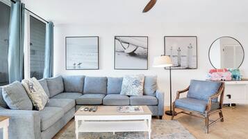 Een woonkamer met een grijze hoekbank, een witte salontafel, een vloerlamp en een houten stoel. Er hangen drie ingelijste foto's aan de muur.