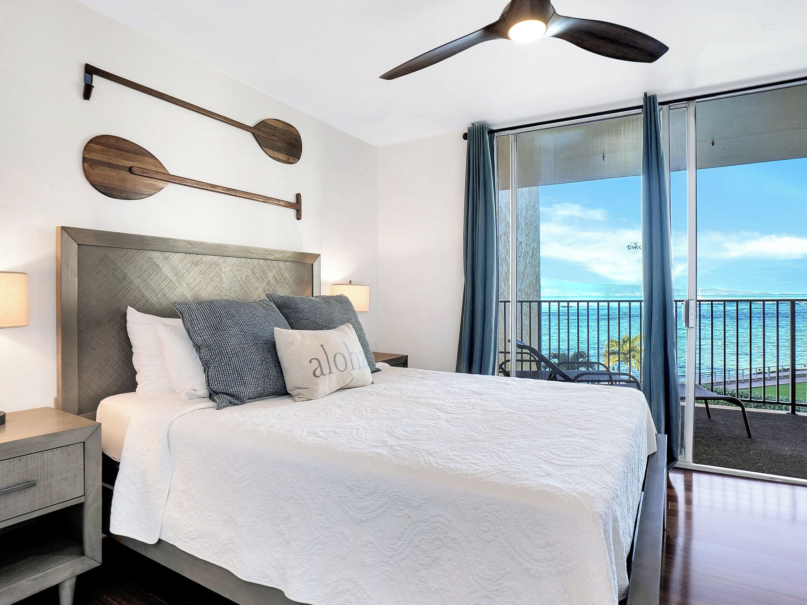 A hotel room with a bed, a nightstand, a wall-mounted lamp, a ceiling fan, and a view of the ocean through a large window.