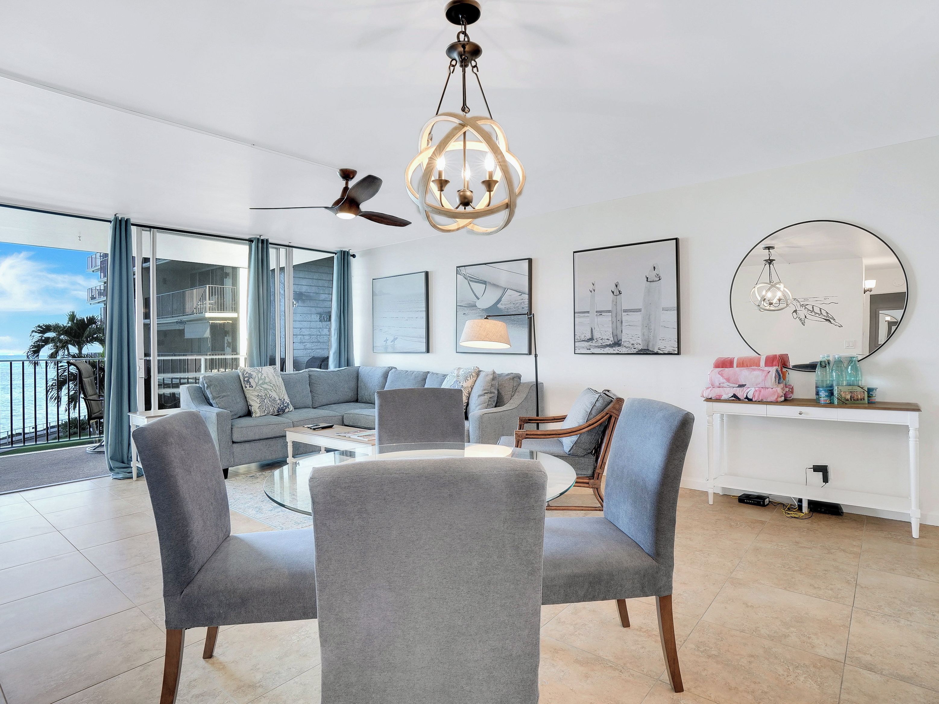 A modern living room with a sectional sofa, a round coffee table, and a dining area with chairs. There is a balcony with a view of the ocean, a ceiling fan, and a chandelier.