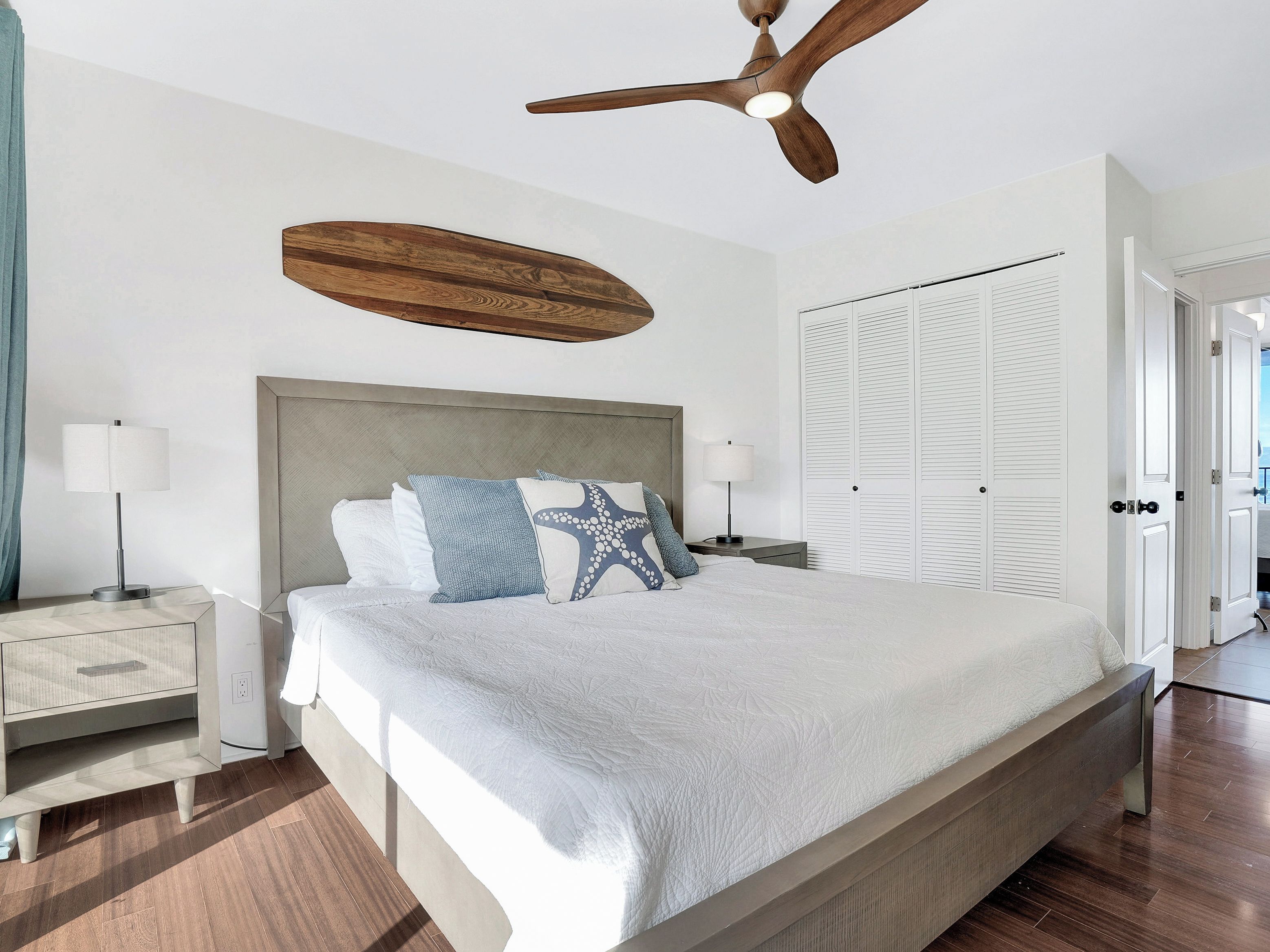 A bedroom with a bed, bedside tables, a ceiling fan, a wardrobe, and a view of the outside.