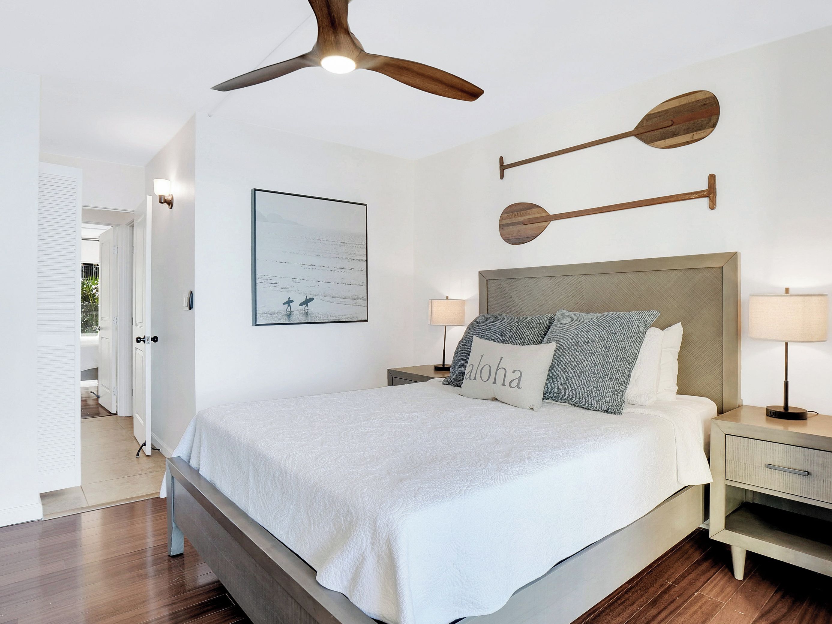 Une chambre à coucher comprenant un lit, des tables de chevet, un ventilateur de plafond, une œuvre d’art accrochée au mur et une porte donnant sur une autre pièce.