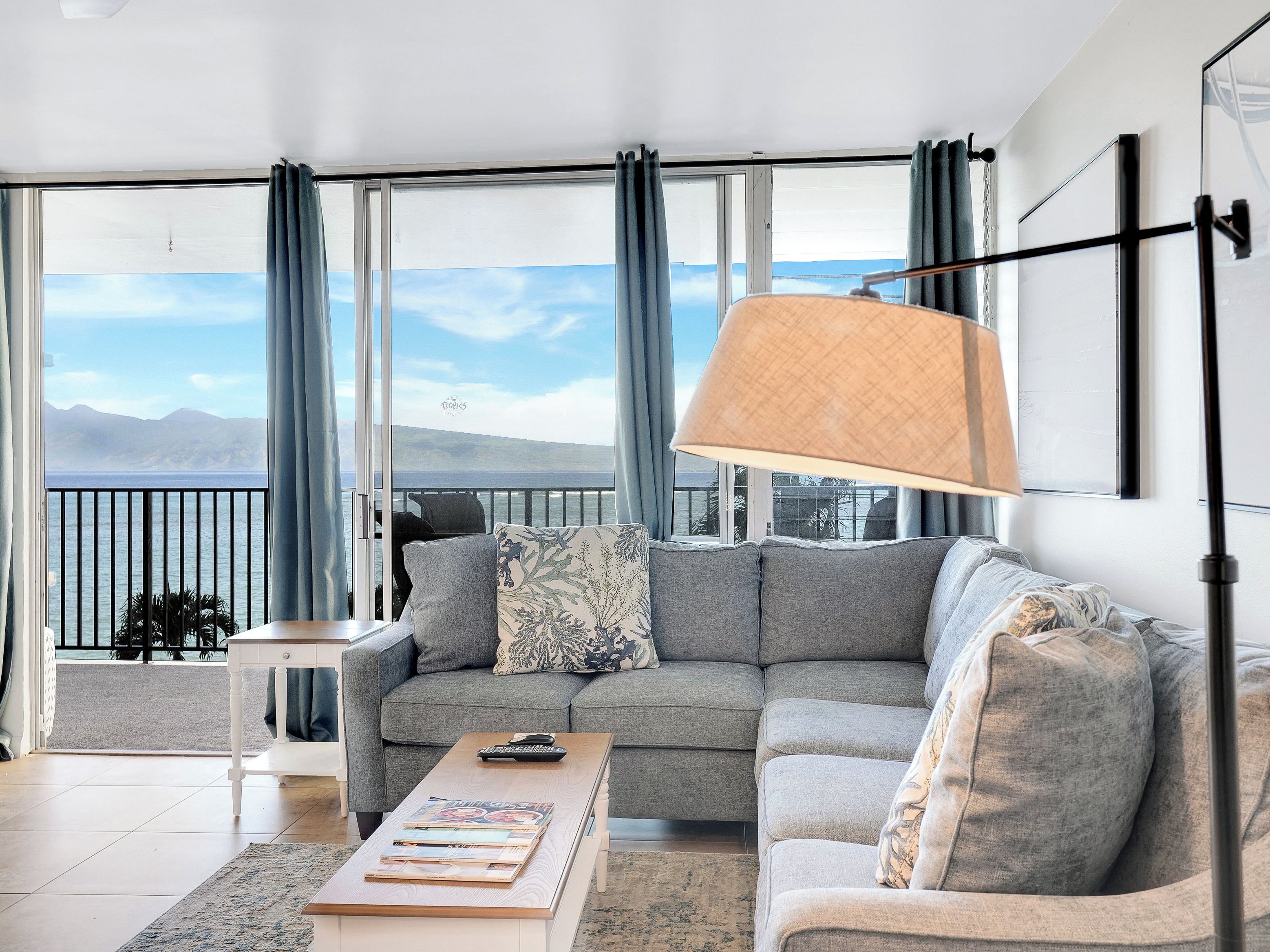 A modern living room with a gray sofa, a coffee table with magazines, a lamp, and a view of the sea through large windows.