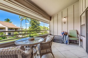 A balcony with wicker furniture, a glass table, and a view of palm trees and houses. - Relax in Kihei | Ocean View Condo, Pool, Hot Tub, Walk to Beach (Kihei)